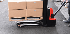 Electric pallet jack lifting a wooden pallet stacked with cardboard boxes on a warehouse floor.