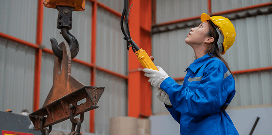 A worker wearing safety attire using a controller to activate a hook hoisting a heavy industrial item