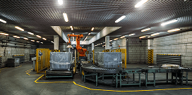 An orange manipulator crane moves a large package on a conveyor system in an industrial facility