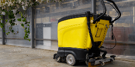A yellow floor cleaning machine is parked in an office hallway in front of a partition and hanging plants