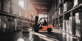 Forklift operator moving palletized goods inside a large warehouse with tall storage racks and natural light.